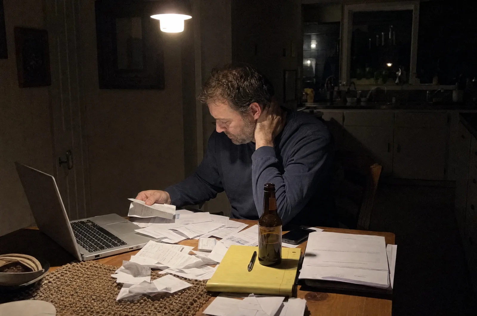 A contractor at his kitchen table at night, doing invoices under a single warm light
